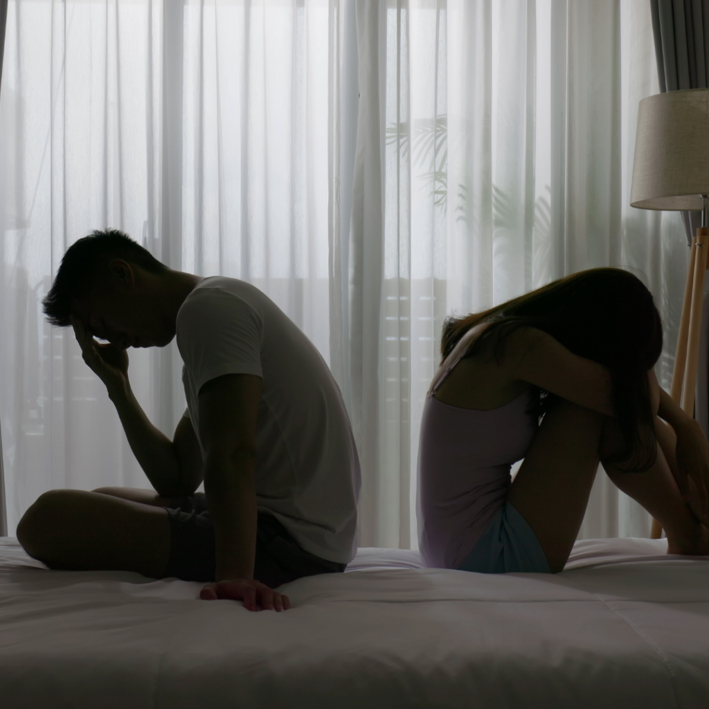 A man and a woman sit back-to-back on a bed in silhouetted postures of distress, evoking the emotional struggle of compulsive sexual behavior disorder.
