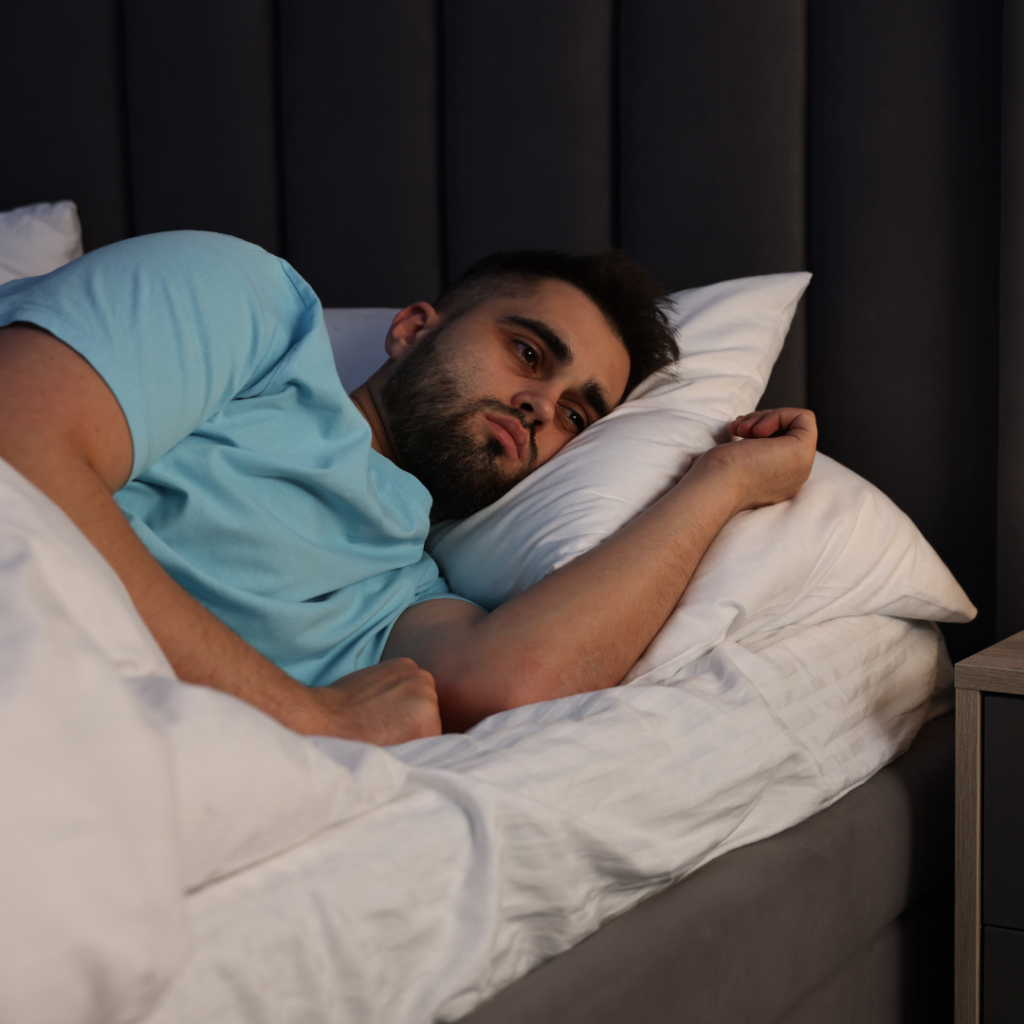 A bearded man in a blue t-shirt lies awake in bed, looking to the side as he reflects on his sex addiction therapy.