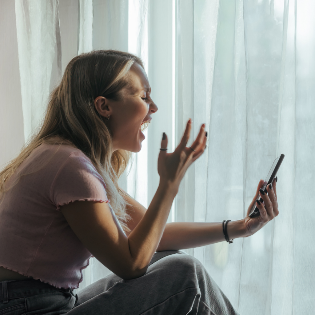 A woman sits by a window, shouting angrily into a smartphone during a virtual anger management therapy session.