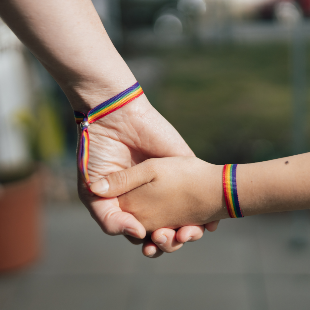 An adult hand and a child’s hand holding each other, both wearing rainbow-striped fabric bracelets symbolizing LGBTQIA mental health support.